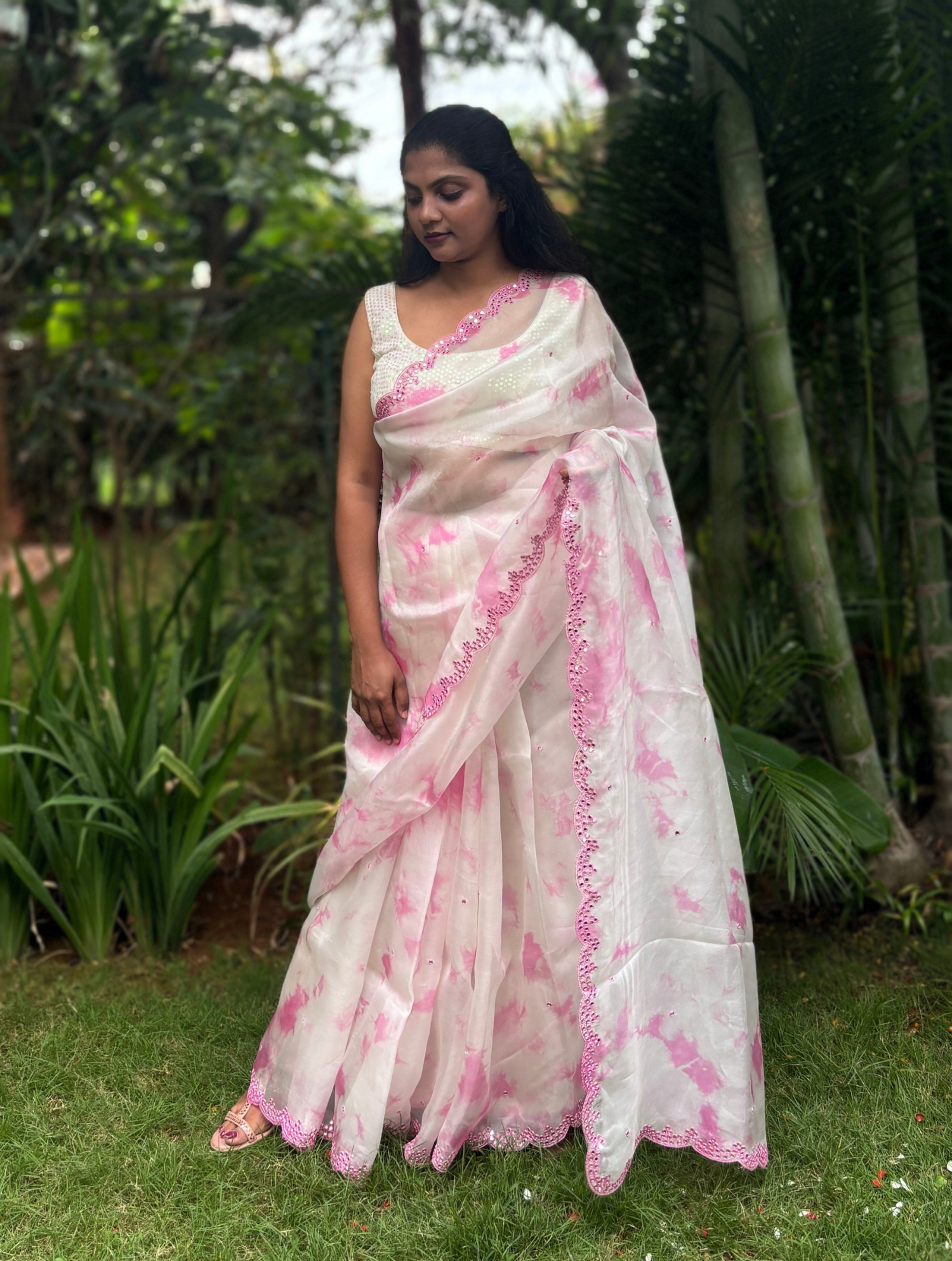 A woman wearing a white and pink tie-dye saree with pink border trim, standing outdoors on a grassy lawn with tropical plants and trees in the background.