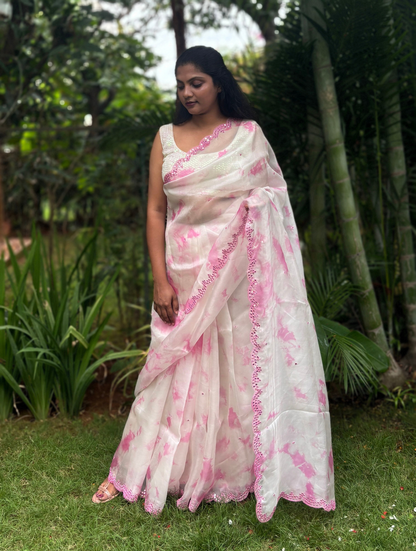 A woman wearing a white and pink tie-dye saree with pink border trim, standing outdoors on a grassy lawn with tropical plants and trees in the background.