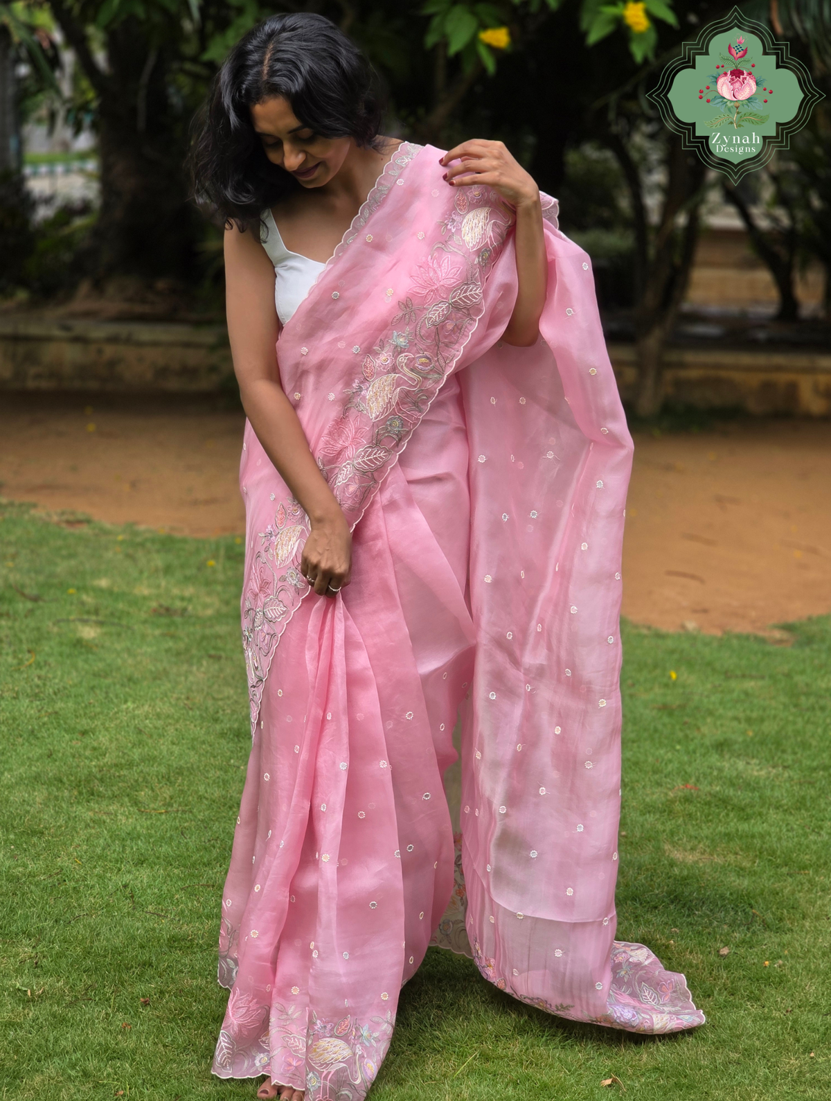 A woman wearing a pink organza silk saree with all over small embroidered butis and crane and floral embroidery on the borders, standing outdoors on a green lawn with trees in the background. 