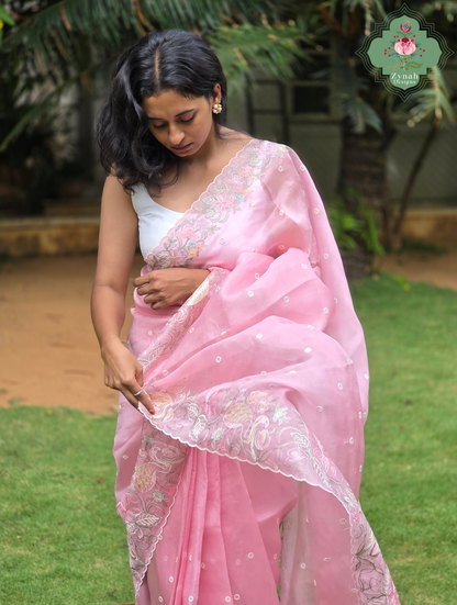A woman wearing a pink organza saree with crane & floral embroidery and a white sleeveless blouse and the pallu in her hand, standing outdoors on a grassy lawn with trees in the background.