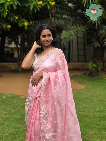 A woman wearing a pink organza silk saree with crane & floral embroidery, standing outdoors in a garden setting with green grass and trees in the background.